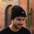 A man wearing a black knit beanie with a white embroidered Hidalgo logo, photographed outdoors in front of a warm-toned building with a cactus in the background.