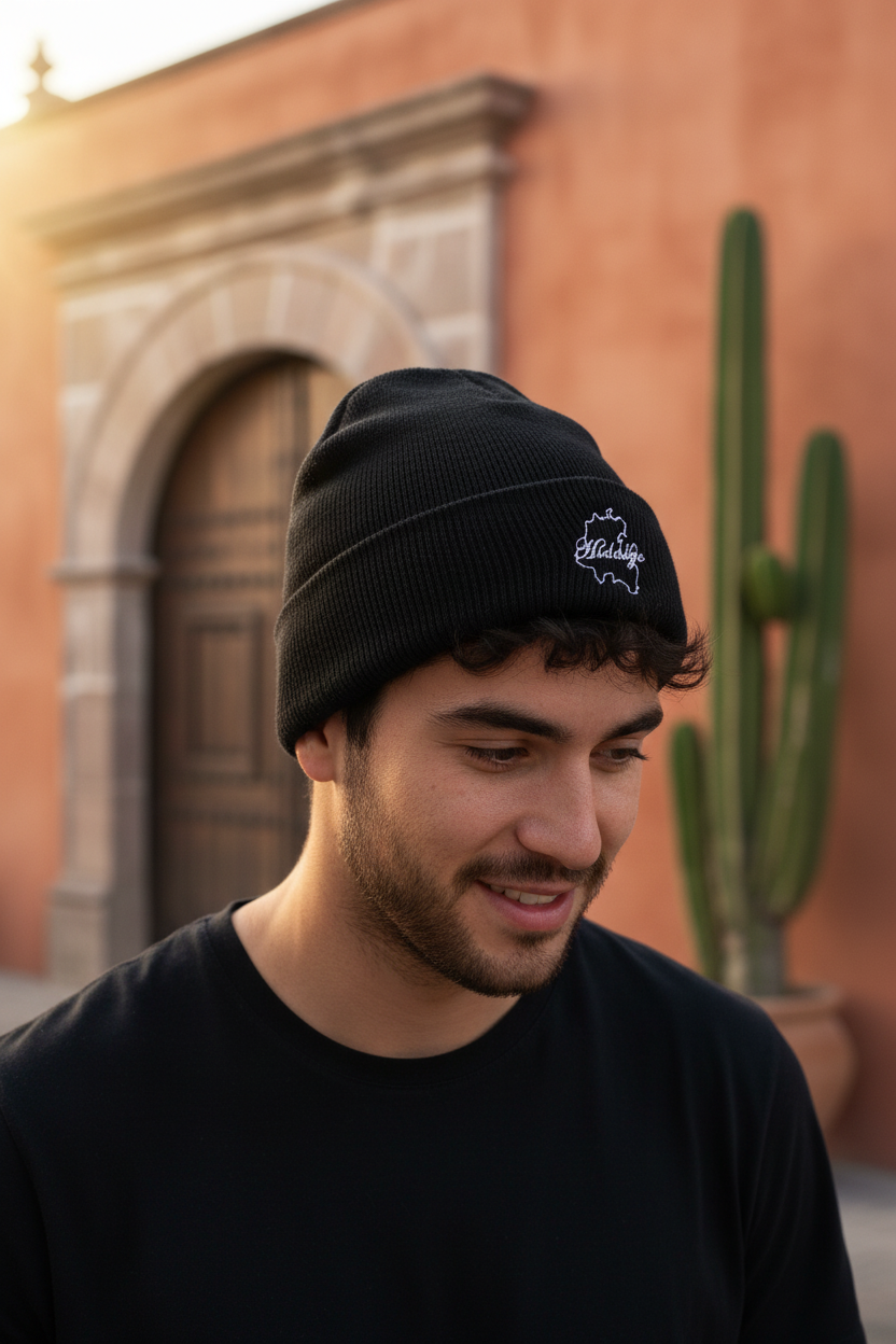 A man wearing a black knit beanie with a white embroidered Hidalgo logo, photographed outdoors in front of a warm-toned building with a cactus in the background.