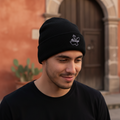 A man wearing a black knit beanie with a small white Jalisco embroidered logo on the front, standing outdoors in front of a warm-toned stone and brick building with a wooden door.