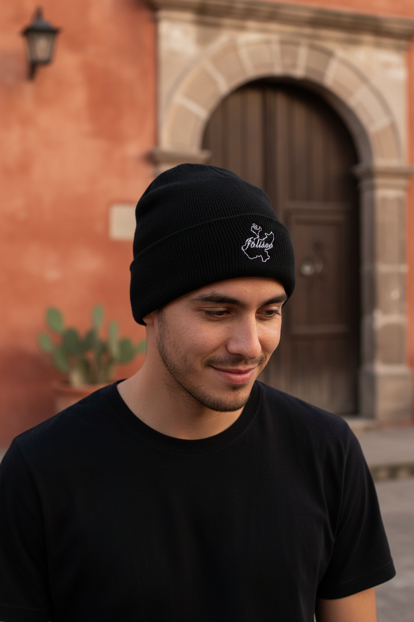 A man wearing a black knit beanie with a small white Jalisco embroidered logo on the front, standing outdoors in front of a warm-toned stone and brick building with a wooden door.