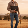 A man wearing a tan overshirt, blue jeans, brown boots, and sunglasses stands in a dry grass field at sunset, holding a black leather clutch bag.