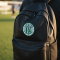 A black backpack worn by a person on a grassy field, featuring a circular green and white Leon FC crest on the front pocket.