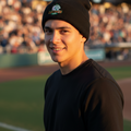 A smiling man wearing a black knit beanie with a small multicolor 'Leones de Yucatan' logo and a black t-shirt, standing on a baseball field with a blurred stadium crowd in the background.