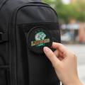 A close-up of a black backpack with a circular embroidered patch featuring a horse head logo and the text 'LEONES VICTOR' being held by a hand. The background is a blurred outdoor street scene.