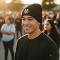 A smiling young man outdoors at sunset wearing a black Mariachis de Guadalajara knit beanie with a white stylized logo and a black long-sleeve shirt, with blurred people in the background.