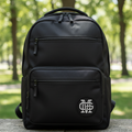 A black backpack with a silver interlocking 'Mariachis de Guadalajara' logo patch on the front, resting on a stone ledge with a blurred green park background.