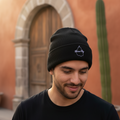 A man wearing a black Mexico City knit beanie with a small white geometric logo on the front, standing outdoors in front of a warm terracotta-colored building with a wooden door and a tall cactus.