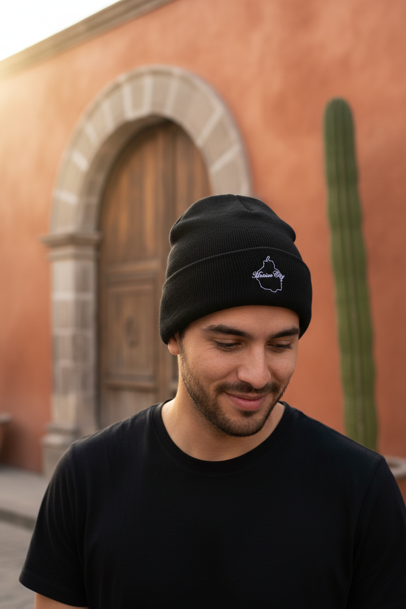 A man wearing a black Mexico City knit beanie with a small white geometric logo on the front, standing outdoors in front of a warm terracotta-colored building with a wooden door and a tall cactus.