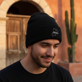 A man wearing a black Mexico knit beanie with a small white embroidered logo, standing outdoors in front of a warm-toned stucco building with an arched doorway and a cactus.