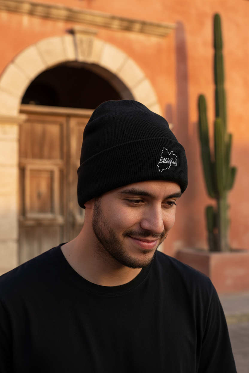 A man wearing a black Mexico knit beanie with a small white embroidered logo, standing outdoors in front of a warm-toned stucco building with an arched doorway and a cactus.
