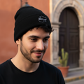 A man wearing a black Michoacan knit beanie with a white embroidered logo, standing outdoors in front of a warm-toned building with a stone archway.