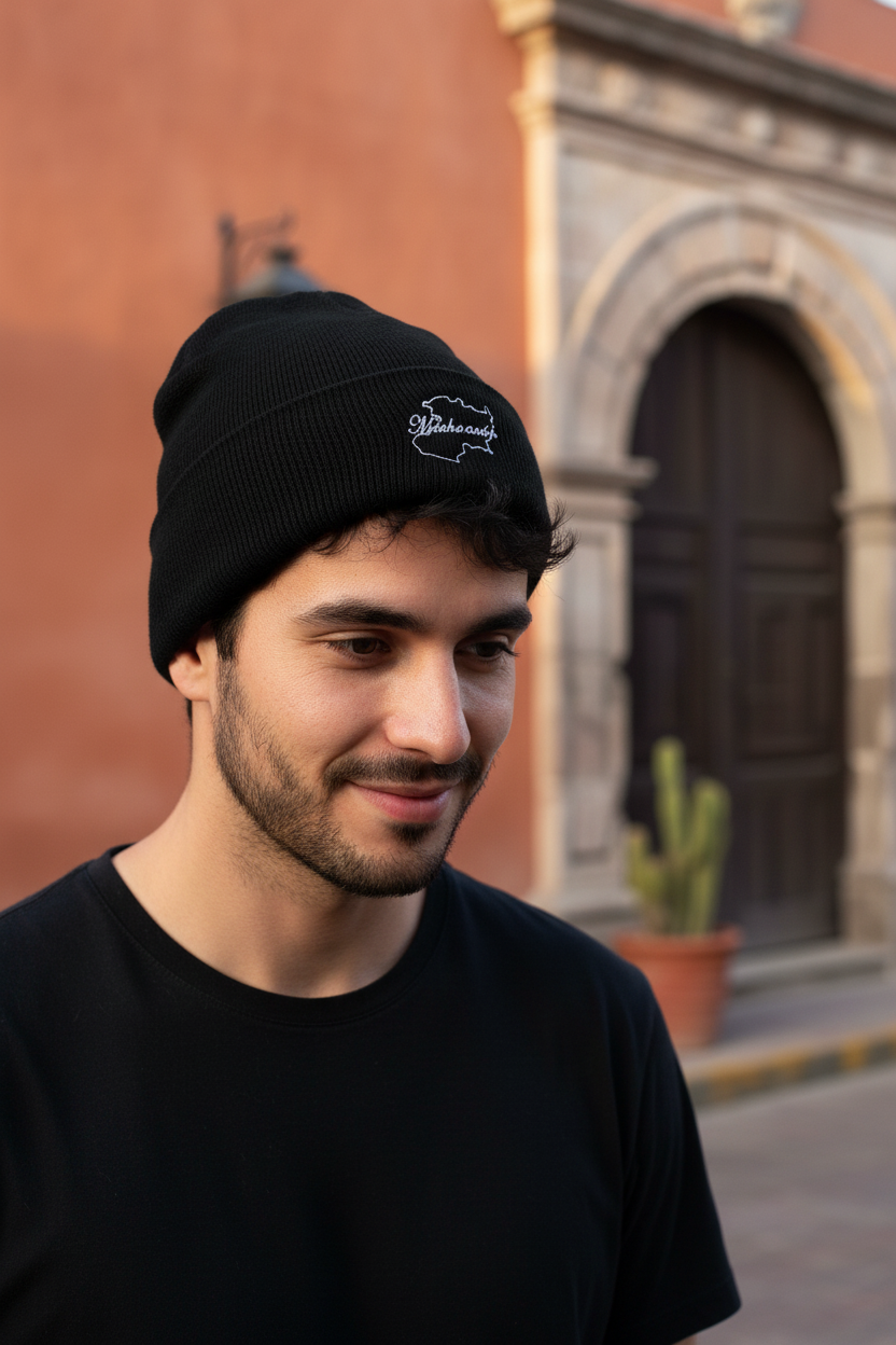 A man wearing a black Michoacan knit beanie with a white embroidered logo, standing outdoors in front of a warm-toned building with a stone archway.