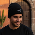 A man wearing a black Morelos knit beanie with a white embroidered logo, standing outdoors in front of a warm-toned stone building with a wooden door.