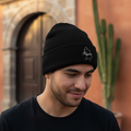 A man wearing a black Nayarit knit beanie with a small white embroidered logo on the front, standing outdoors in front of a warm-toned building with a cactus.