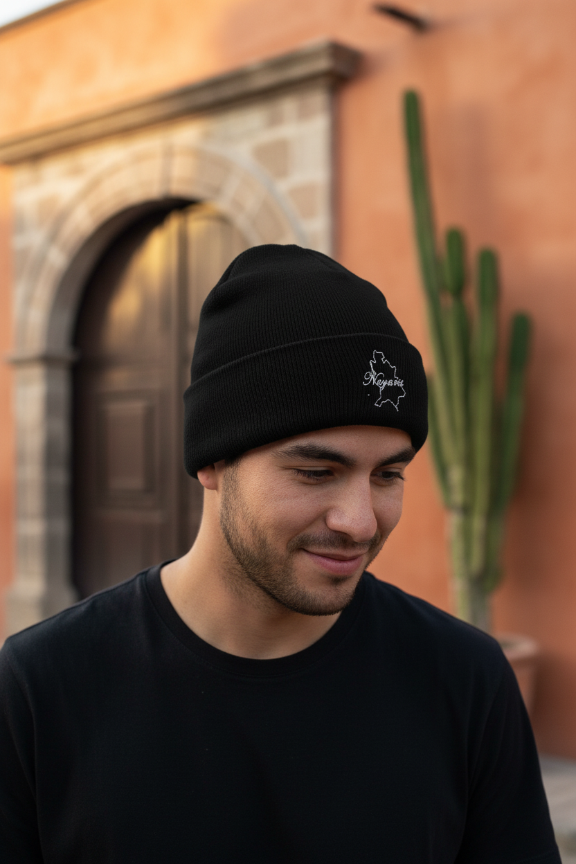 A man wearing a black Nayarit knit beanie with a small white embroidered logo on the front, standing outdoors in front of a warm-toned building with a cactus.