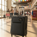 A black rolling suitcase with an extended telescoping handle stands in a brightly lit airport terminal. The suitcase has a front zippered pocket, a circular black-and-white logo patch on the upper front, and spinner wheels.