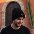 A man wearing a black NUevo Leon knit beanie with a small white embroidered logo on the front, standing outdoors in front of a warm terracotta-colored building with a wooden door and a tall cactus.