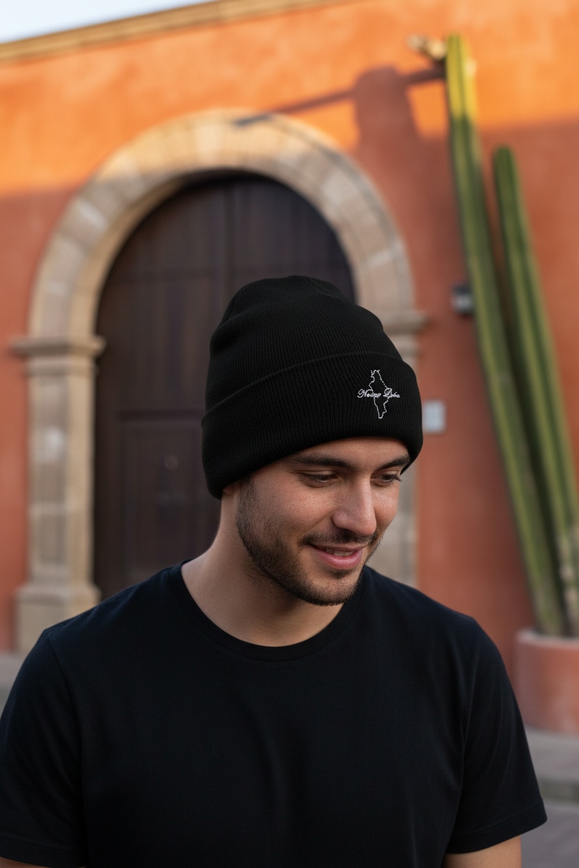 A man wearing a black NUevo Leon knit beanie with a small white embroidered logo on the front, standing outdoors in front of a warm terracotta-colored building with a wooden door and a tall cactus.
