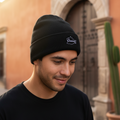 A man wearing a black Oaxaca knit beanie with a white embroidered logo that reads 'Chivas' in an outdoor setting with a warm-toned building and cactus in the background.