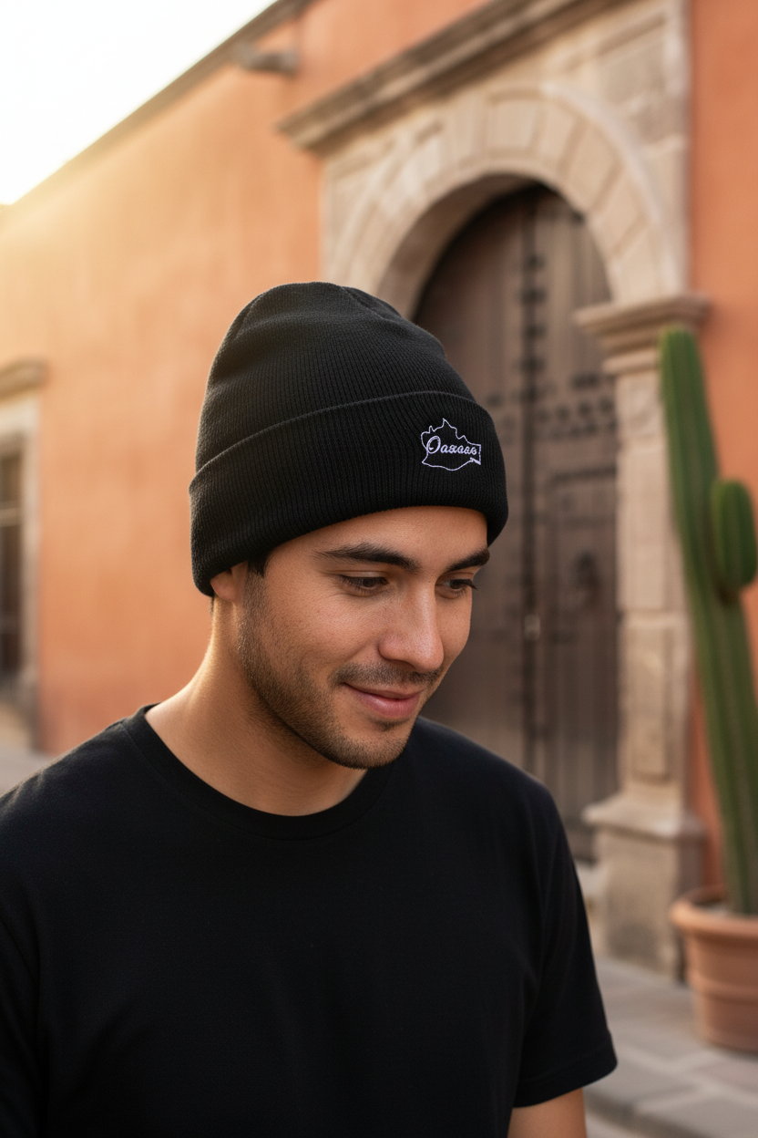 A man wearing a black Oaxaca knit beanie with a white embroidered logo that reads 'Chivas' in an outdoor setting with a warm-toned building and cactus in the background.