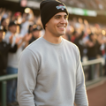 A smiling man wearing a black beanie with a colorful 'Olmecas de Tabasco ' logo and a light gray crewneck sweatshirt, standing on a baseball field with a blurred crowd in the stadium background.