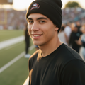 A smiling young man wearing a black knit beanie with a Piratas de Campeche logo and a black t-shirt, standing on a sports field at sunset.