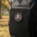 A close-up of a black backpack being held outdoors, with a circular front patch reading “Pirates” and a bird logo. A person’s hand and part of their body are visible at the top, and the background is a blurred park setting.
