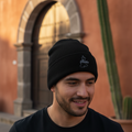 A man wearing a black Puebla knit beanie with a small white embroidered logo, standing outdoors in front of a warm-toned stucco building with an arched doorway and a tall cactus.