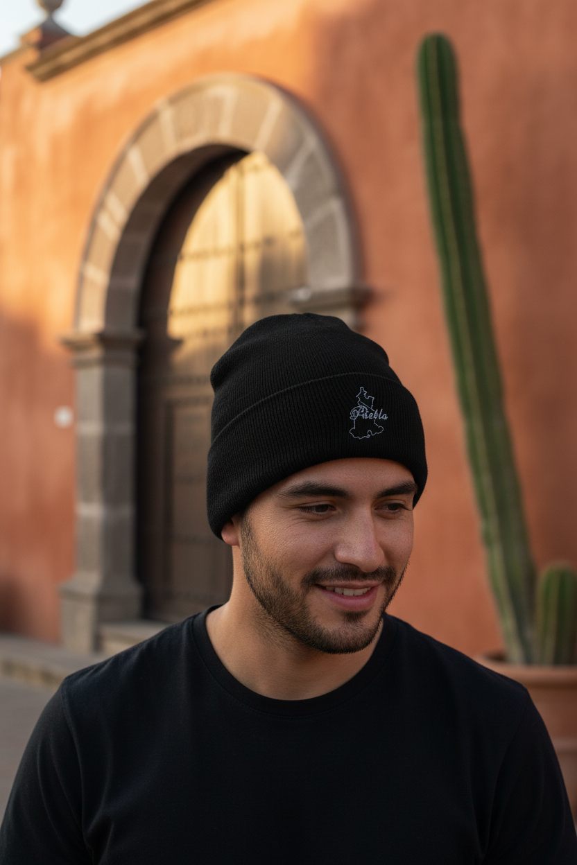 A man wearing a black Puebla knit beanie with a small white embroidered logo, standing outdoors in front of a warm-toned stucco building with an arched doorway and a tall cactus.