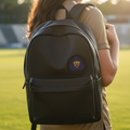 A woman on a grassy field at sunset wearing a black backpack with a purple and gold crest Pumas UNAM logo patch on the front.