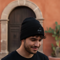 A man wearing a black Queretaro knit beanie with a small white embroidered logo, standing in front of a warm terracotta-colored stucco wall with a stone-trimmed wooden door.