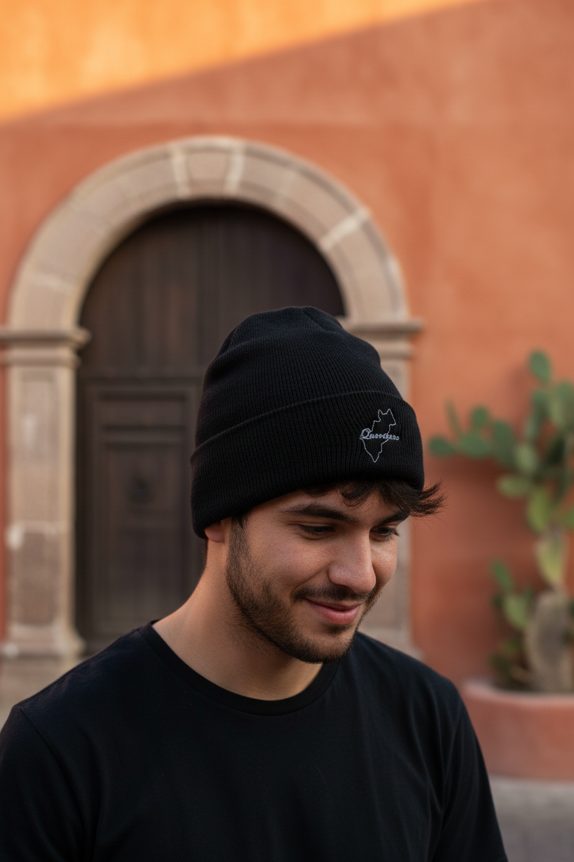 A man wearing a black Queretaro knit beanie with a small white embroidered logo, standing in front of a warm terracotta-colored stucco wall with a stone-trimmed wooden door.