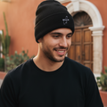 A man wearing a black knit beanie with a small white embroidered logo and a black t-shirt, standing outdoors in front of a warm-toned building.