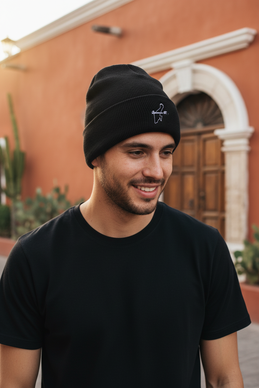 A man wearing a black knit beanie with a small white embroidered logo and a black t-shirt, standing outdoors in front of a warm-toned building.