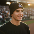 A smiling man in a black Rieleros de Aguascalientes beanie with a white and blue baseball logo and a black t-shirt, standing in a stadium during warm golden hour sunlight.