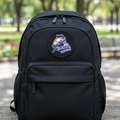 A black backpack with a circular 'Rieleros' patch on the front, sitting on a stone ledge in an outdoor park setting with blurred green trees in the background.