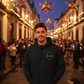 A man wearing a black hoodie with a white outline graphic stands on a cobblestone street at dusk, with people holding candles and colorful hanging star decorations in the background.