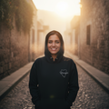 A woman stands smiling in a narrow cobblestone street at sunrise, wearing a black hoodie with a small white chest logo that reads 'Kingsbridge'. The sun is low in the background, creating a warm golden glow and soft haze.