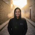 A woman wearing a black hoodie with a small white graphic logo on the left chest stands on a cobblestone street at sunset. The background features warm golden light and blurred stone buildings.
