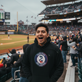 A man wearing a black hoodie with a circular 'Tecolotes Dos Laredos' logo stands in a crowded baseball stadium, laughing and smiling. The background shows the field, stands, and other spectators.