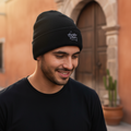 A man wearing a black San Luis Potosi knit beanie with a small white embroidered logo and a black t-shirt, standing outdoors in front of a warm-toned stucco building with a wooden door.