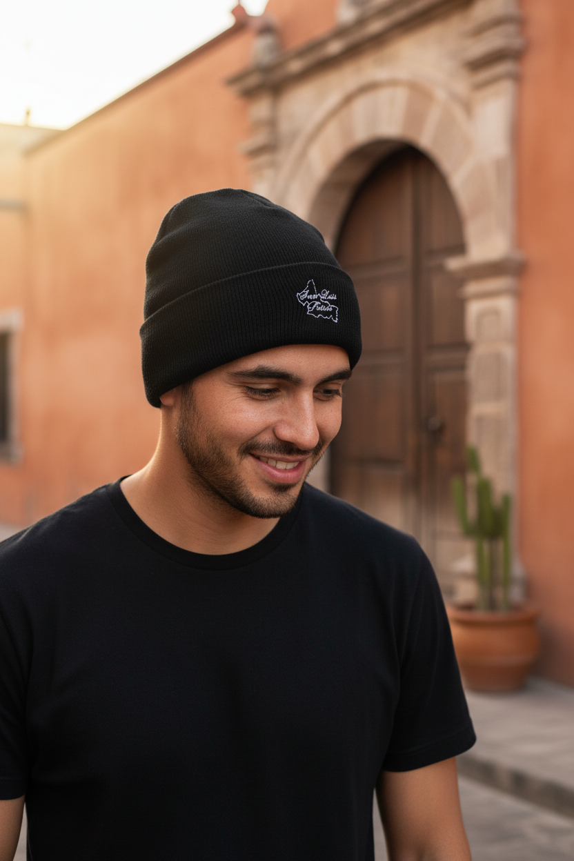 A man wearing a black San Luis Potosi knit beanie with a small white embroidered logo and a black t-shirt, standing outdoors in front of a warm-toned stucco building with a wooden door.