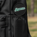 Close-up of a black backpack with a circular black patch featuring the green and white 'Snaperos' script logo, set against a blurred outdoor background.