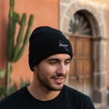 A man wearing a black knit beanie with a white embroidered logo, standing outdoors in front of a warm-toned building with a cactus on the left.