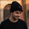 A man wearing a black Sonora knit beanie with a small white embroidered logo on the front, standing outdoors in front of a warm-toned building with a cactus visible in the background.