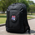 A black backpack with a circular 'Sultanes' patch featuring a red and blue logo, sitting on a stone surface outdoors with a blurred park background.