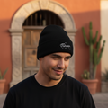 A man wearing a black knit beanie with a white embroidered logo and a black t-shirt, standing outdoors in front of a warm terracotta-colored stucco building with an arched doorway and potted cacti.