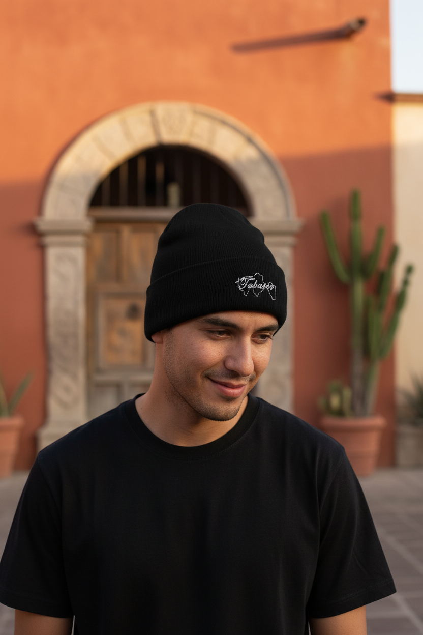 A man wearing a black knit beanie with a white embroidered logo and a black t-shirt, standing outdoors in front of a warm terracotta-colored stucco building with an arched doorway and potted cacti.