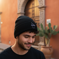 A man wearing a black knit beanie with a small white embroidered Tamaulipas logo, standing outdoors in front of a warm terracotta wall and stone archway.