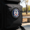 A close-up of a black backpack with a circular embroidered patch reading 'TECOLOTES DOS LAREDOOS' on the front pocket, photographed outdoors with a blurred park background.